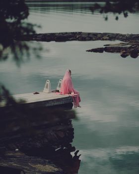 A woman in a pink dress sits peacefully by a serene lakeside, capturing a tranquil moment in nature.