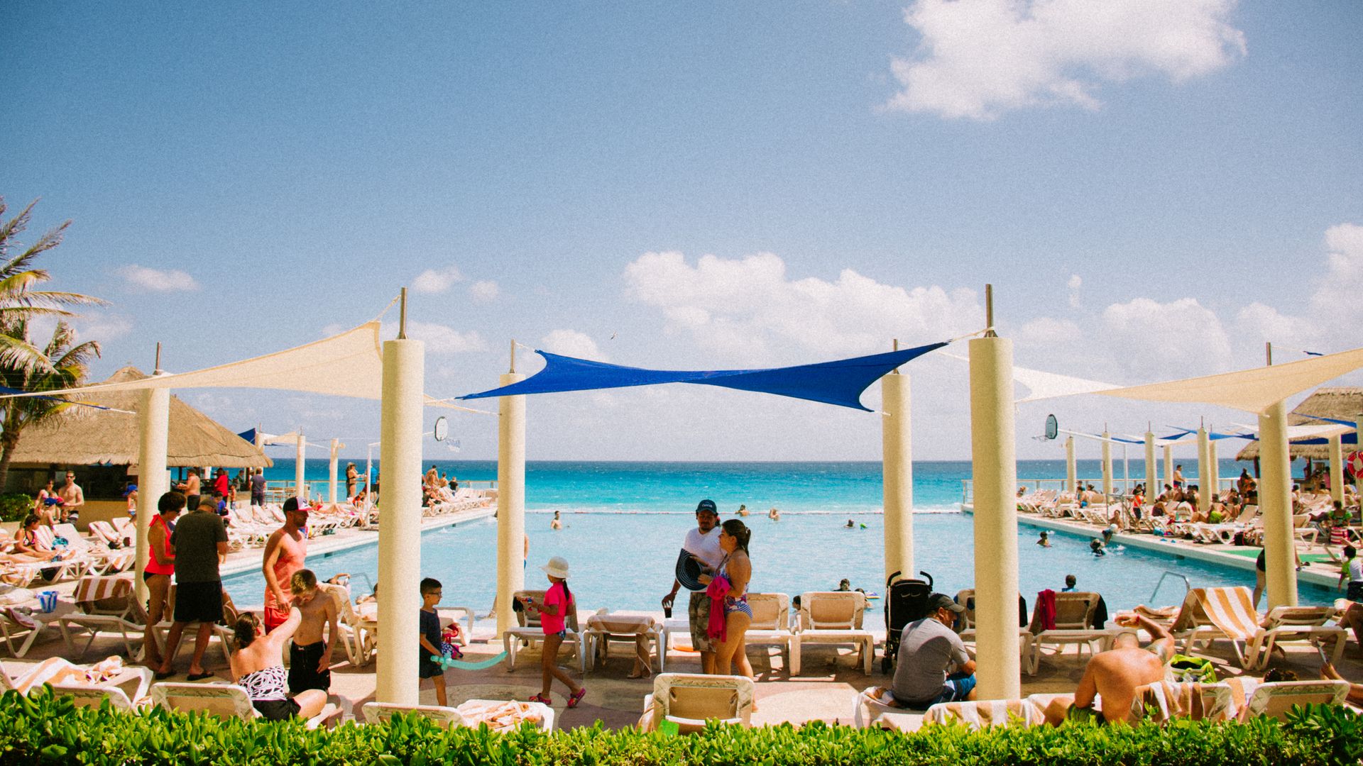 Families enjoy a sunny day at a resort pool overlooking a vibrant blue ocean under a clear sky.