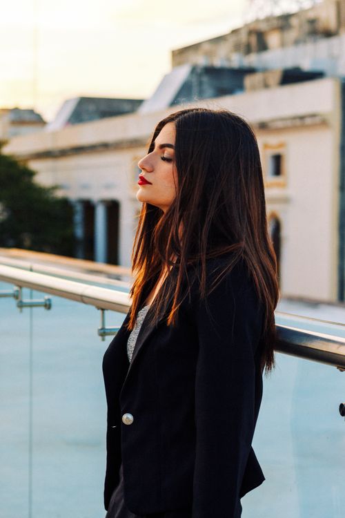 Free Stylish young woman with long brunette hair in a blazer stands on a rooftop railing. Stock Photo