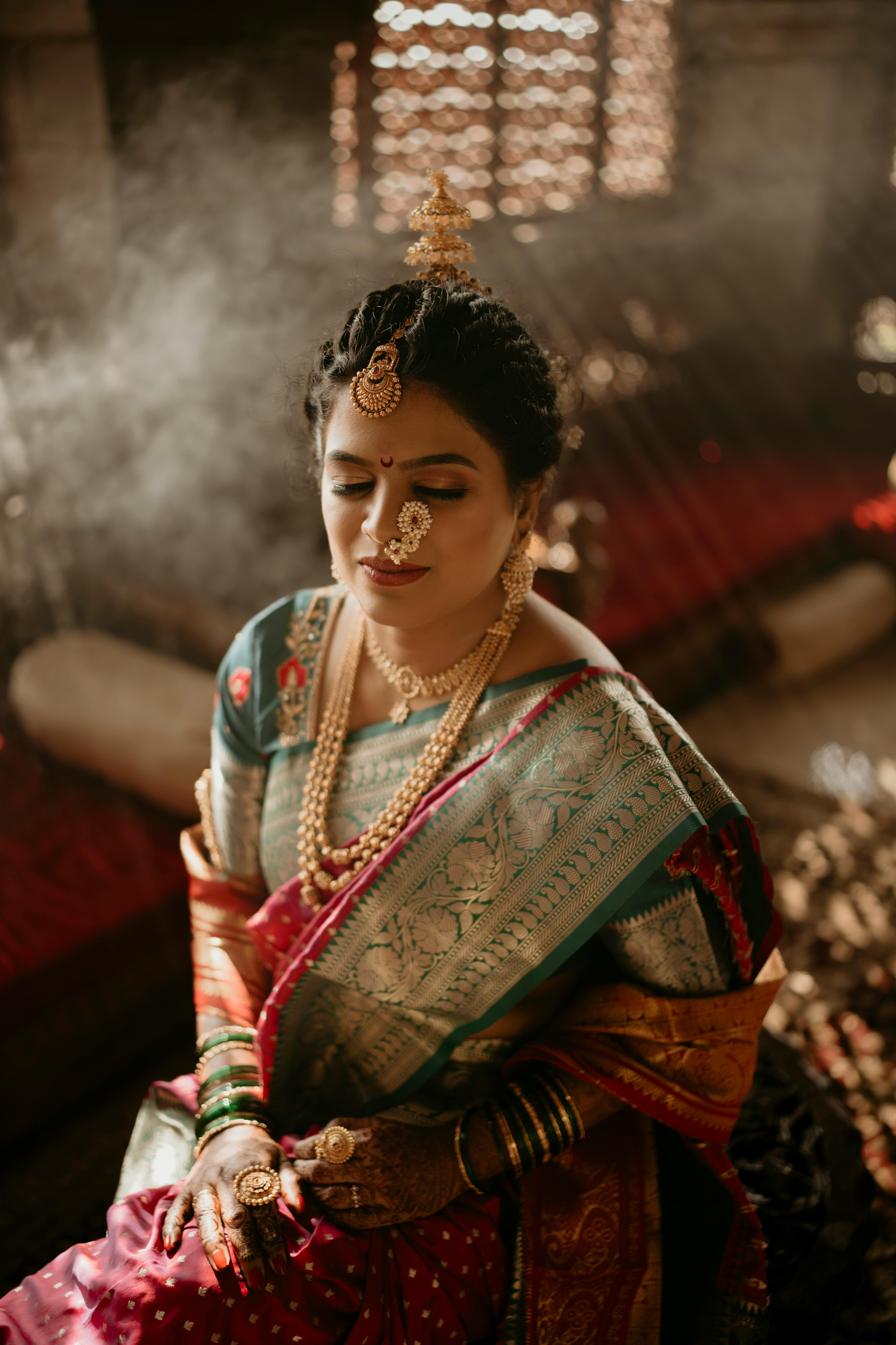 A serene Indian bride wearing jewelry and a saree, sitting indoors with eyes closed, captured in soft sunlight.