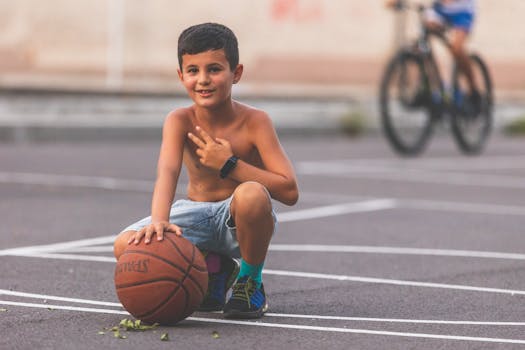 A young boy seated on a basketball court holding a basketball, enjoying a summer day.