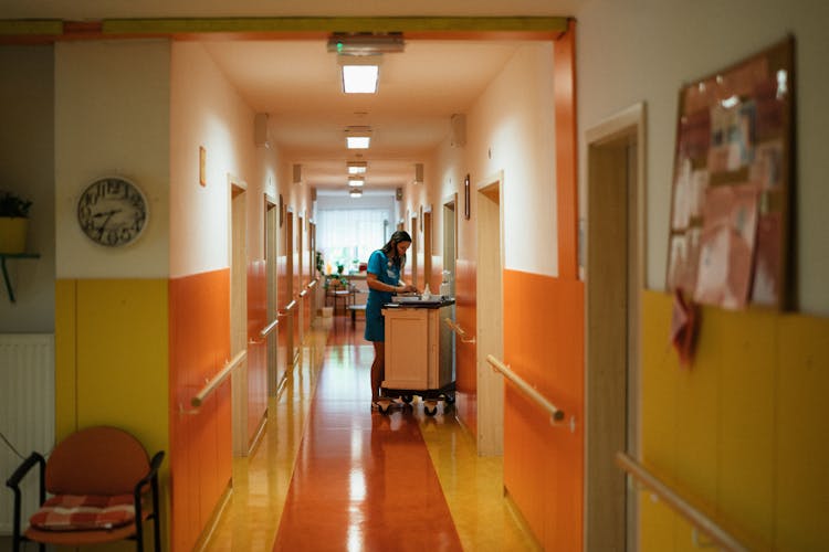 Nurse With Cart On Corridor At Hospital