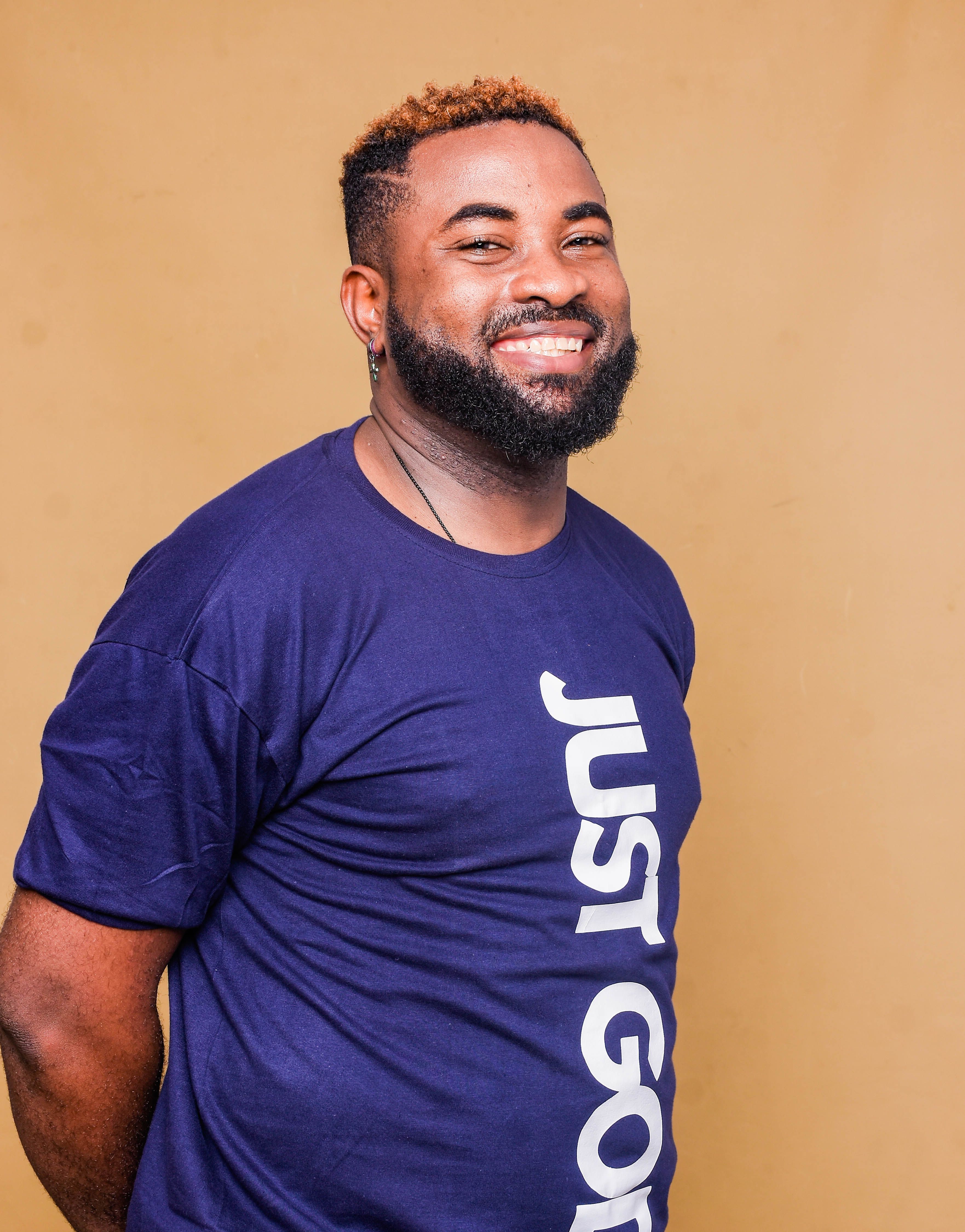 Smiling man in a blue t-shirt posing casually with hands behind back in a studio setting.