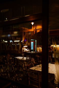 Warmly lit restaurant interior with dining tables and chairs viewed through a window at night.