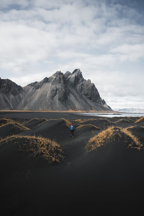Free A lone hiker walks through scenic black sand dunes with a stunning Icelandic mountain backdrop under cloudy skies. Stock Photo