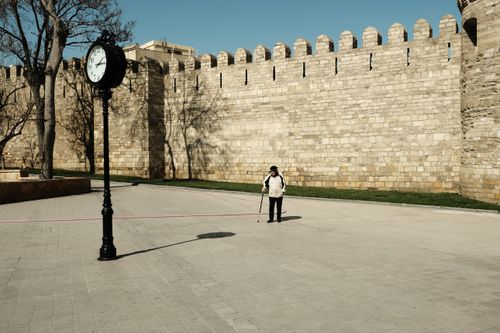 Free An elderly man walks near a historic wall and tower clock on a sunny sidewalk. Stock Photo
