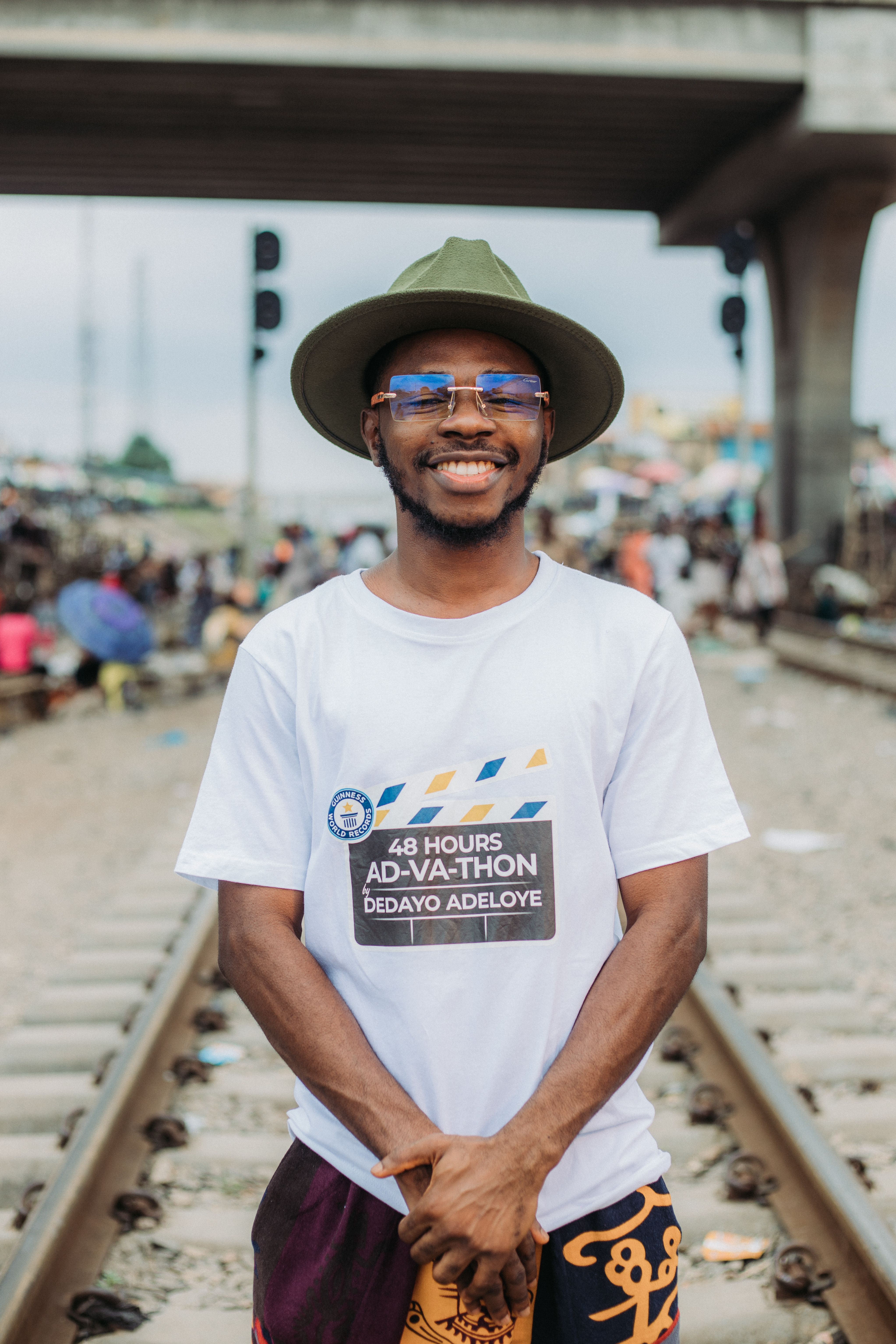 A smiling man in a fedora stands confidently on rail tracks, exuding joy.