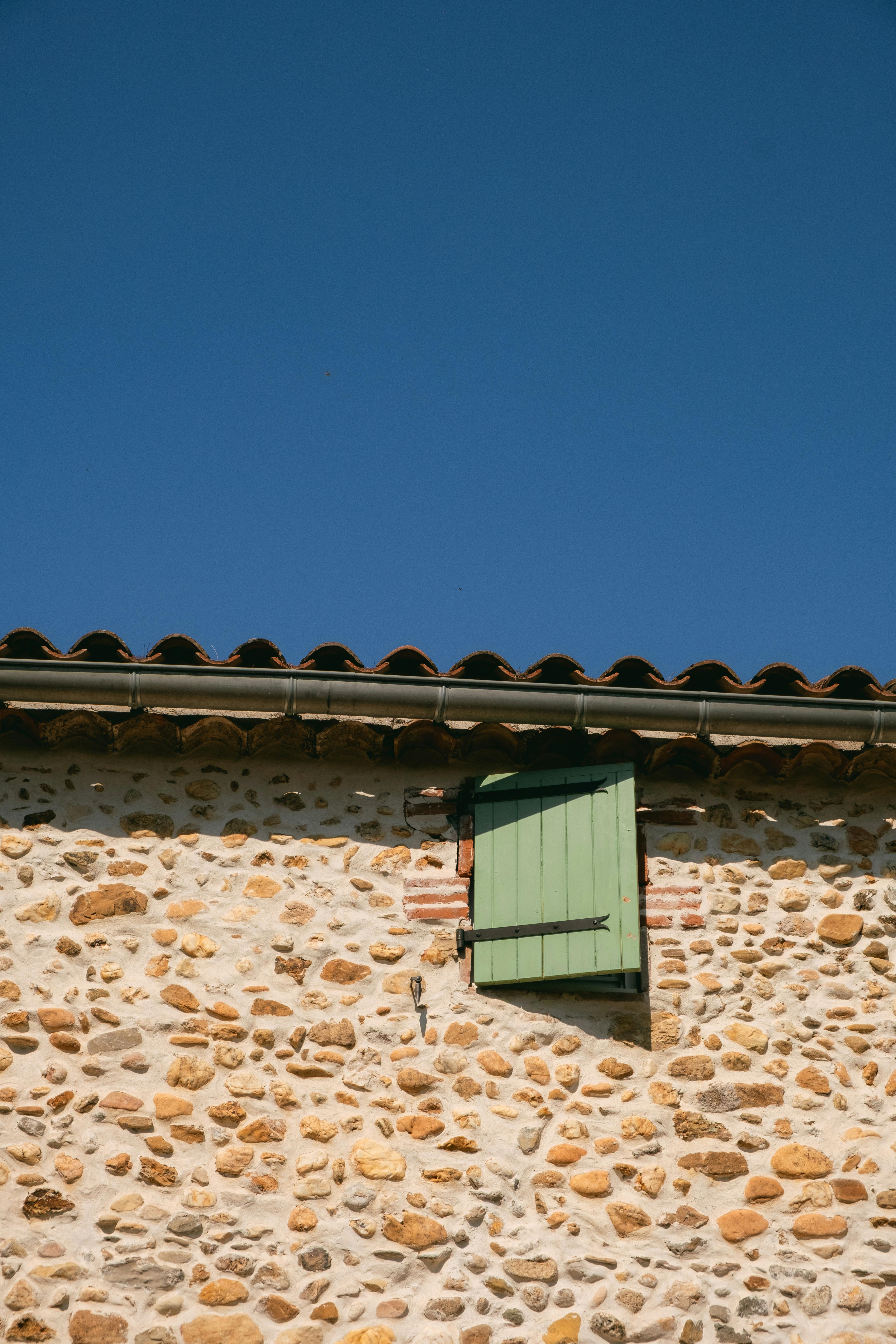 Rustic building exterior with stone wall and green window shutter under a clear blue sky.