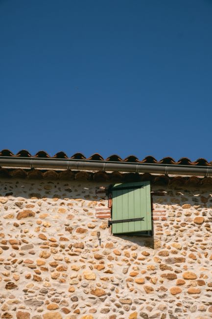 Rustic building exterior with stone wall and green window shutter under a clear blue sky.