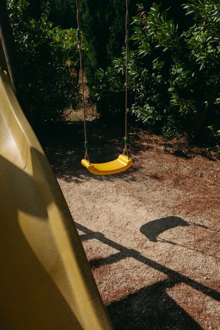 A yellow swing hanging in a sunlit outdoor playground surrounded by greenery.