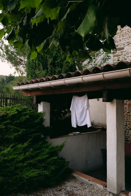 A minimalistic scene of a white shirt drying under a stone-roofed porch amidst greenery.