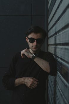 A fashionable man poses against a modern urban backdrop, wearing sunglasses and a smartwatch.
