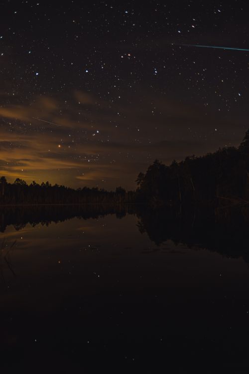 Free A serene night landscape of a starry sky reflecting over a calm lake surrounded by forest. Stock Photo