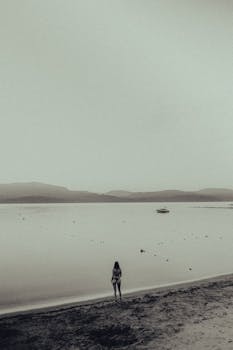 A woman in swimwear stands alone on a misty beach by a serene lake.