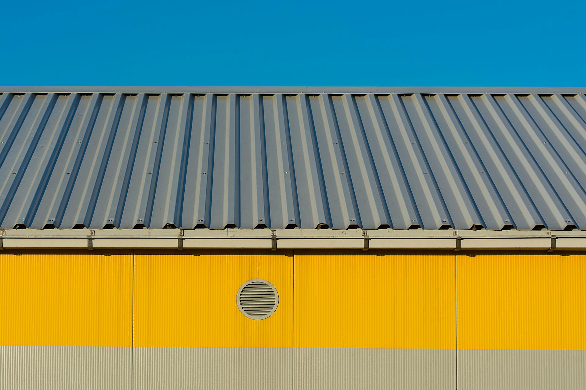 Modern industrial building's yellow facade with metal roof against clear blue sky.