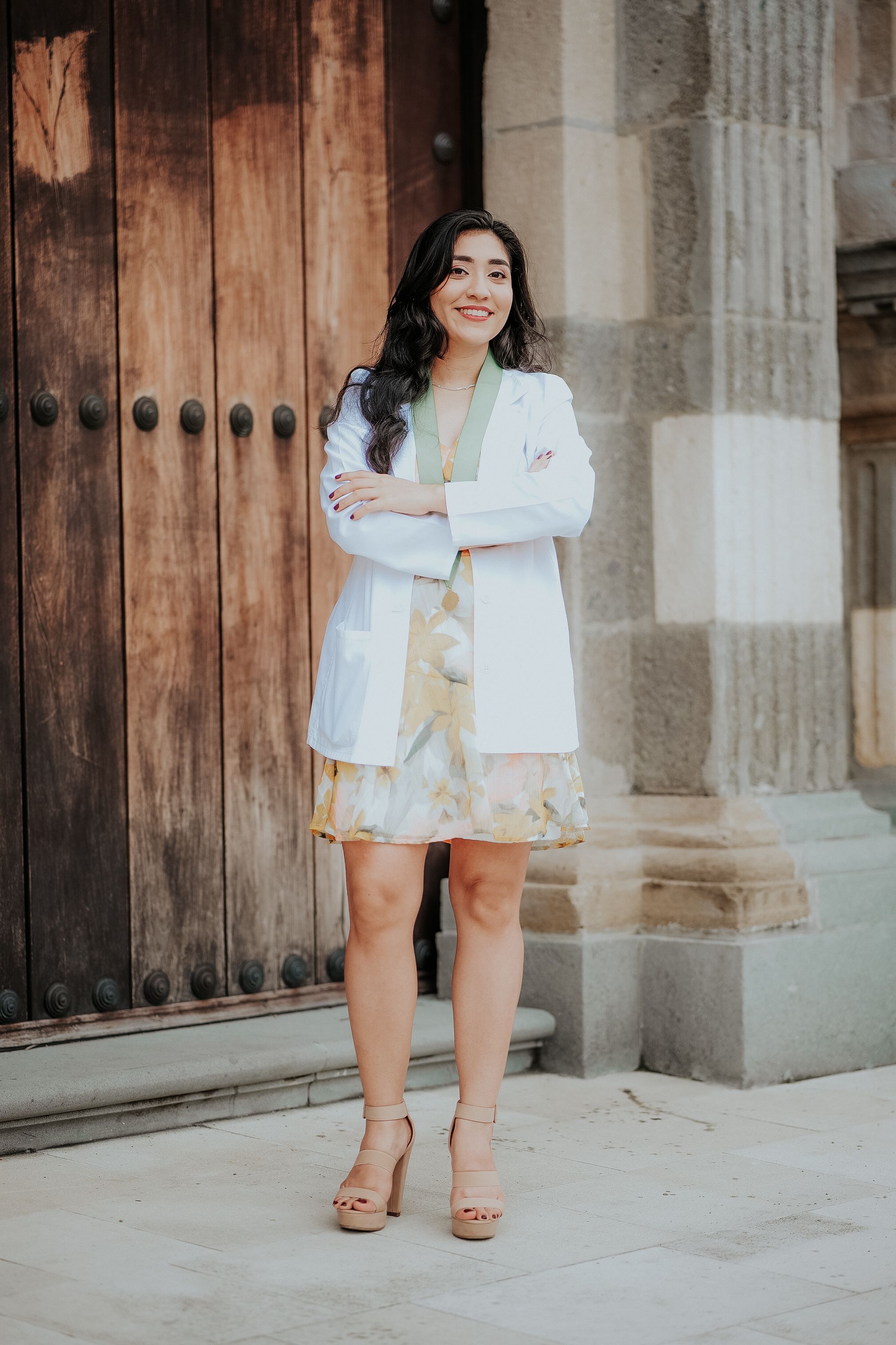 Confident woman in white coat poses by rustic wooden door and ornate pillars.
