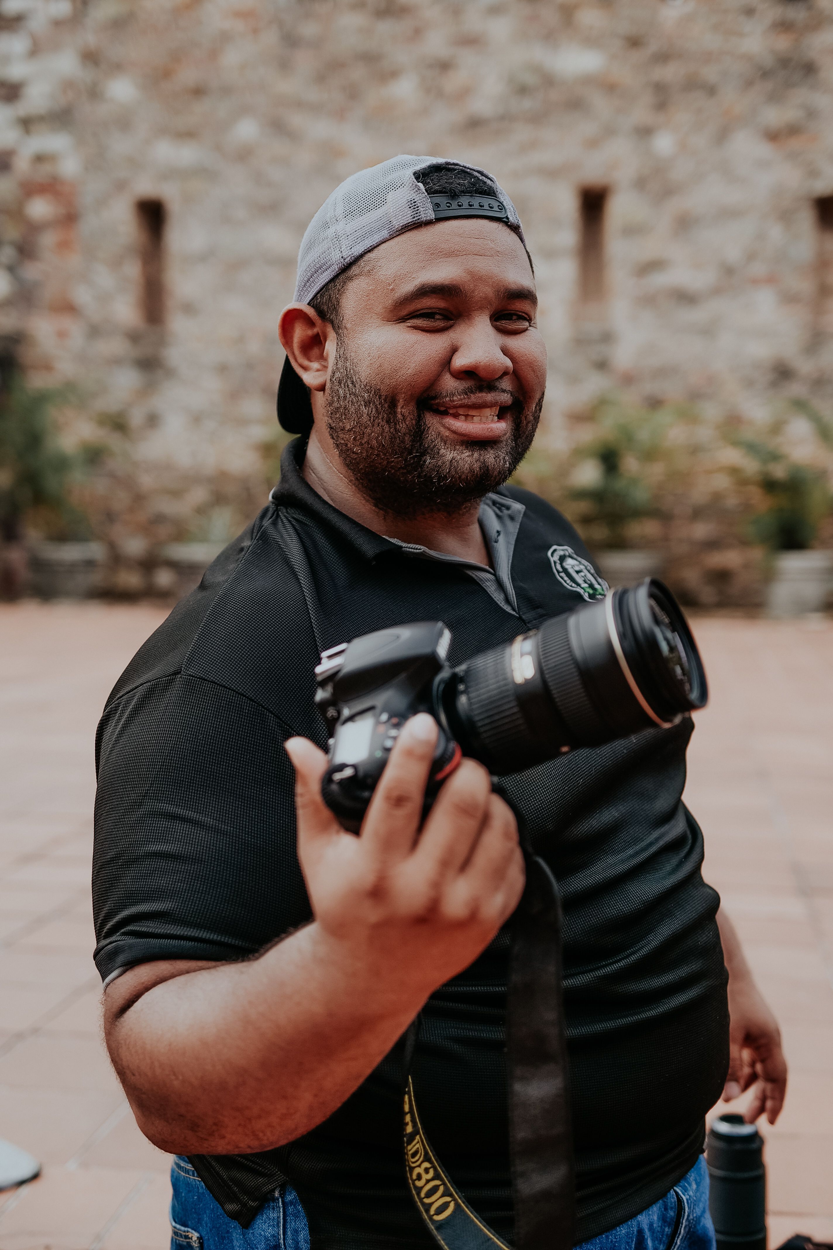 Smiling man holding a camera outdoors, wearing a black t-shirt and cap.