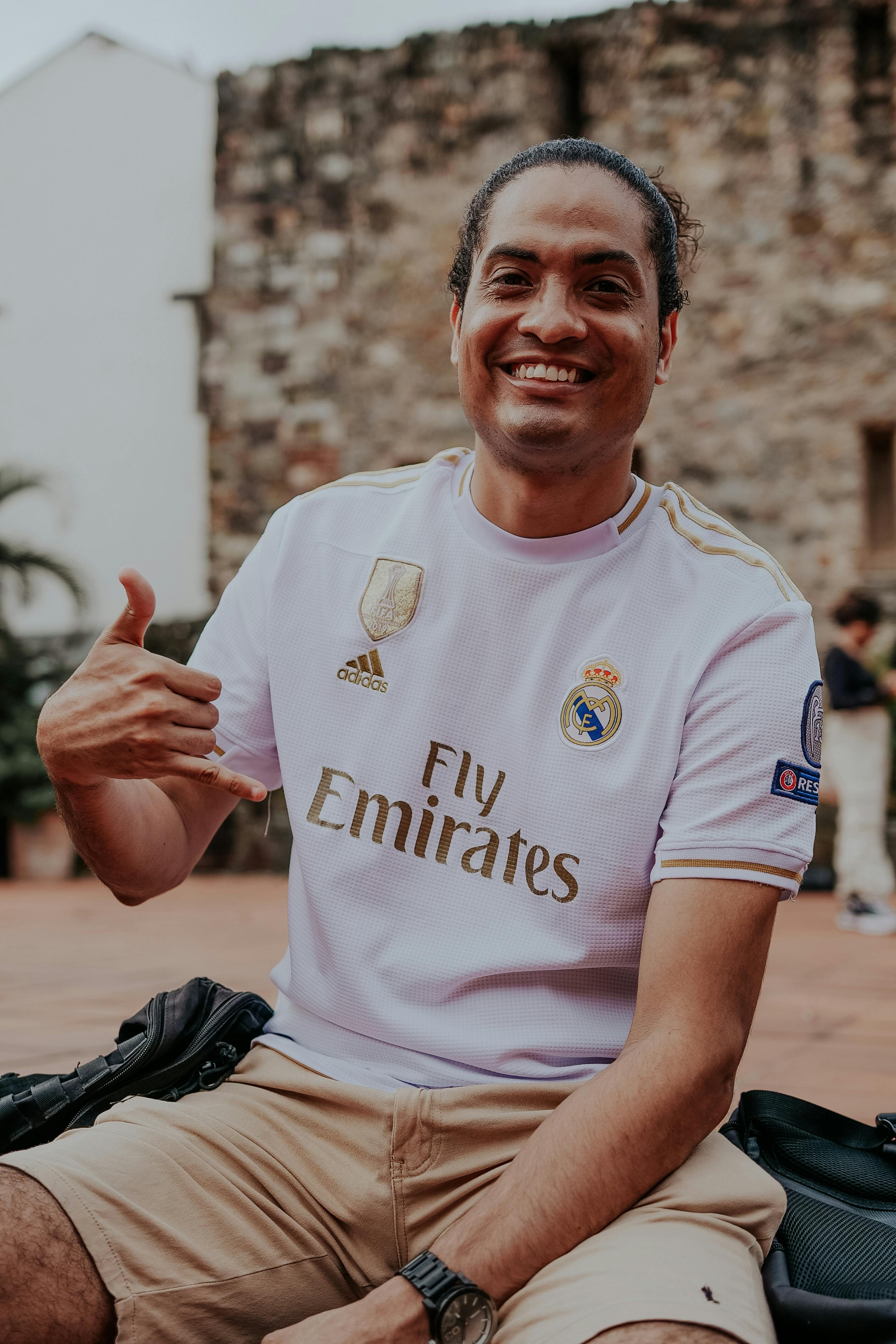 A cheerful man outdoors posing with the shaka sign, wearing a white sports jersey and beige shorts.