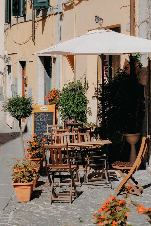 Free Cozy café patio with wooden tables, chairs, and vibrant flowers in a sunlit European city street. Stock Photo