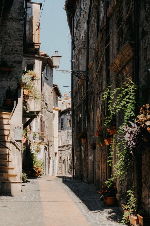 Free Sunlit narrow alleyway with rustic stone buildings and greenery in a historic town street. Stock Photo