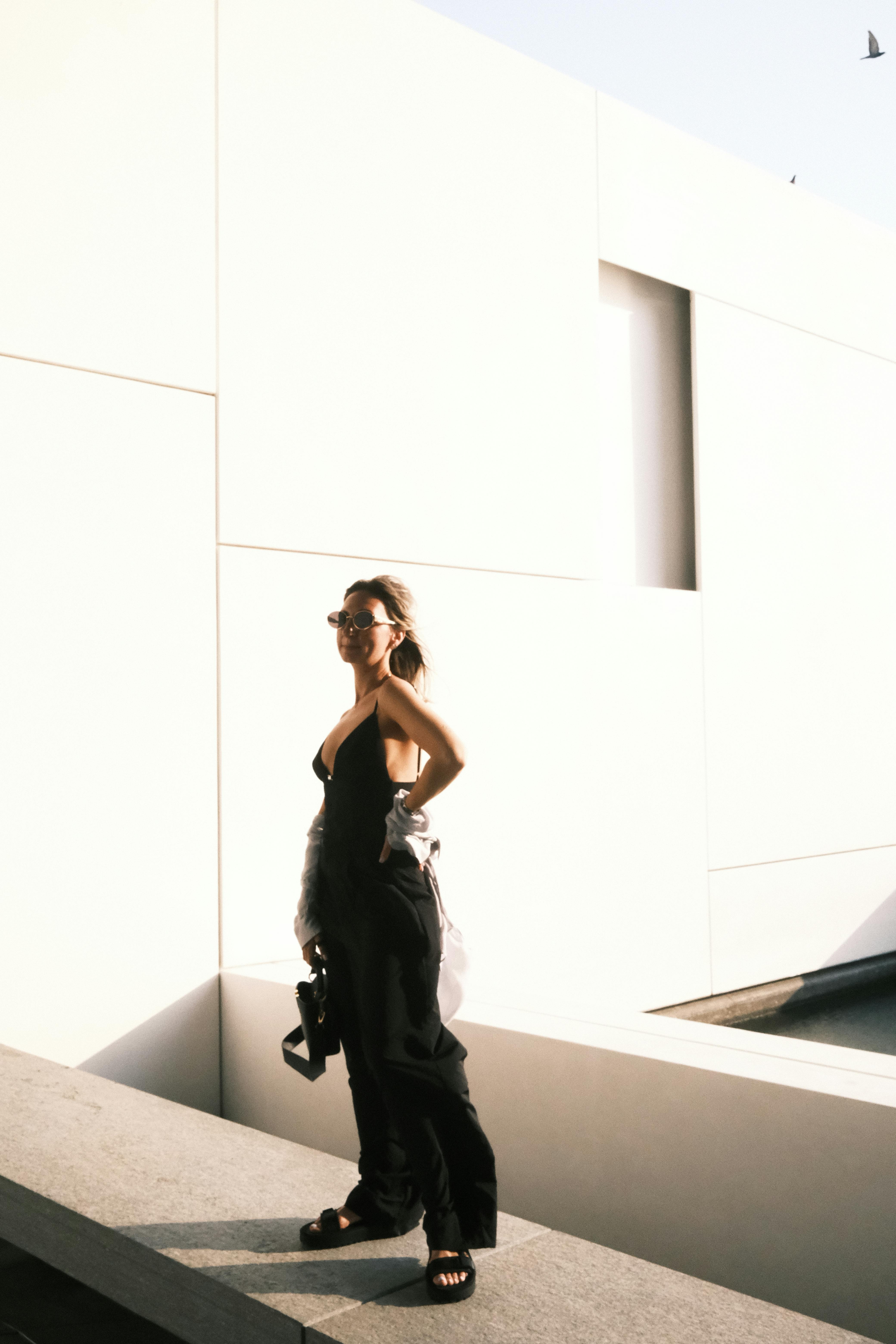 Free A fashionable young woman in a black outfit poses stylishly outdoors against a modern white building. Stock Photo