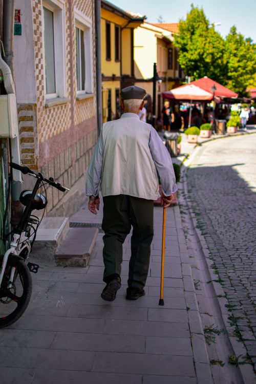 Free Back view of an elderly man walking with a cane on a quaint city sidewalk, embracing urban life. Stock Photo