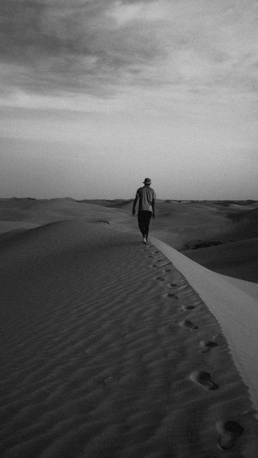 Black and white image of a man walking on sand dunes in Algeria, leaving footprints.