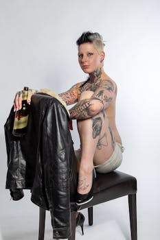Portrait of a tattooed woman sitting on a stool with a leather jacket and drink in a studio setting.
