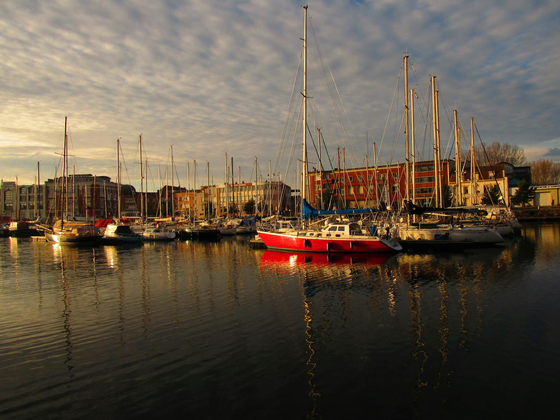Peaceful harbor scene at Dunkerque with sailboats moored at sunset reflecting on calm water.