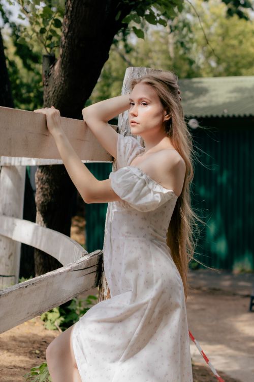 Free A woman in a white dress poses elegantly against a wooden fence in a rural setting. Stock Photo