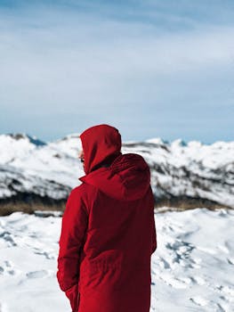 A lone hiker in a red jacket traverses a snowy mountain landscape during a winter day.