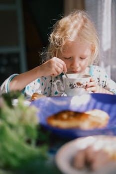 A young blonde girl enjoys a breakfast of tea and pastries in soft morning light.