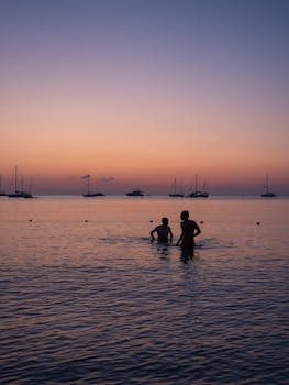 Bathers enjoy a serene sunset by the sea with silhouetted boats in the distance.