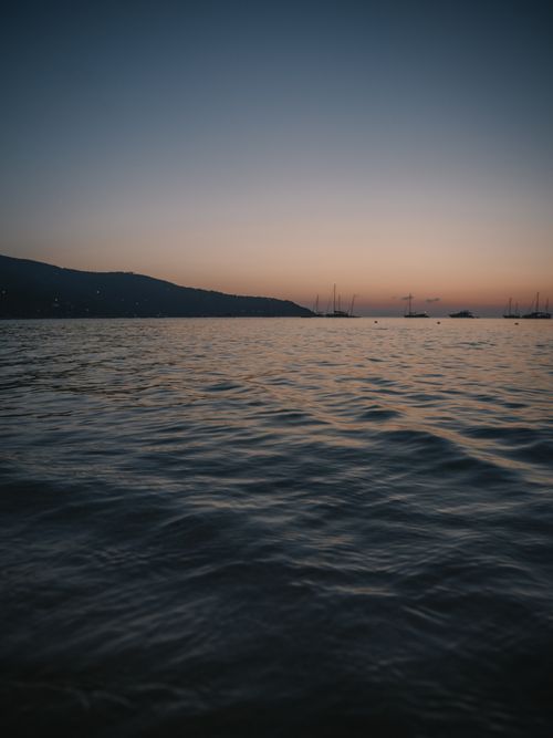 Free A serene coastal scene with sailboats silhouetted against a warm sunset sky and calm waters. Stock Photo