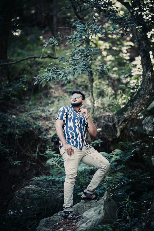 Free A fashionable man in a patterned shirt poses confidently amidst lush forest greenery. Stock Photo