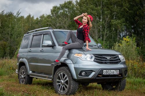 Free A woman sitting on a 4x4 SUV in a rural meadow, smiling with scenic forest backdrop. Stock Photo
