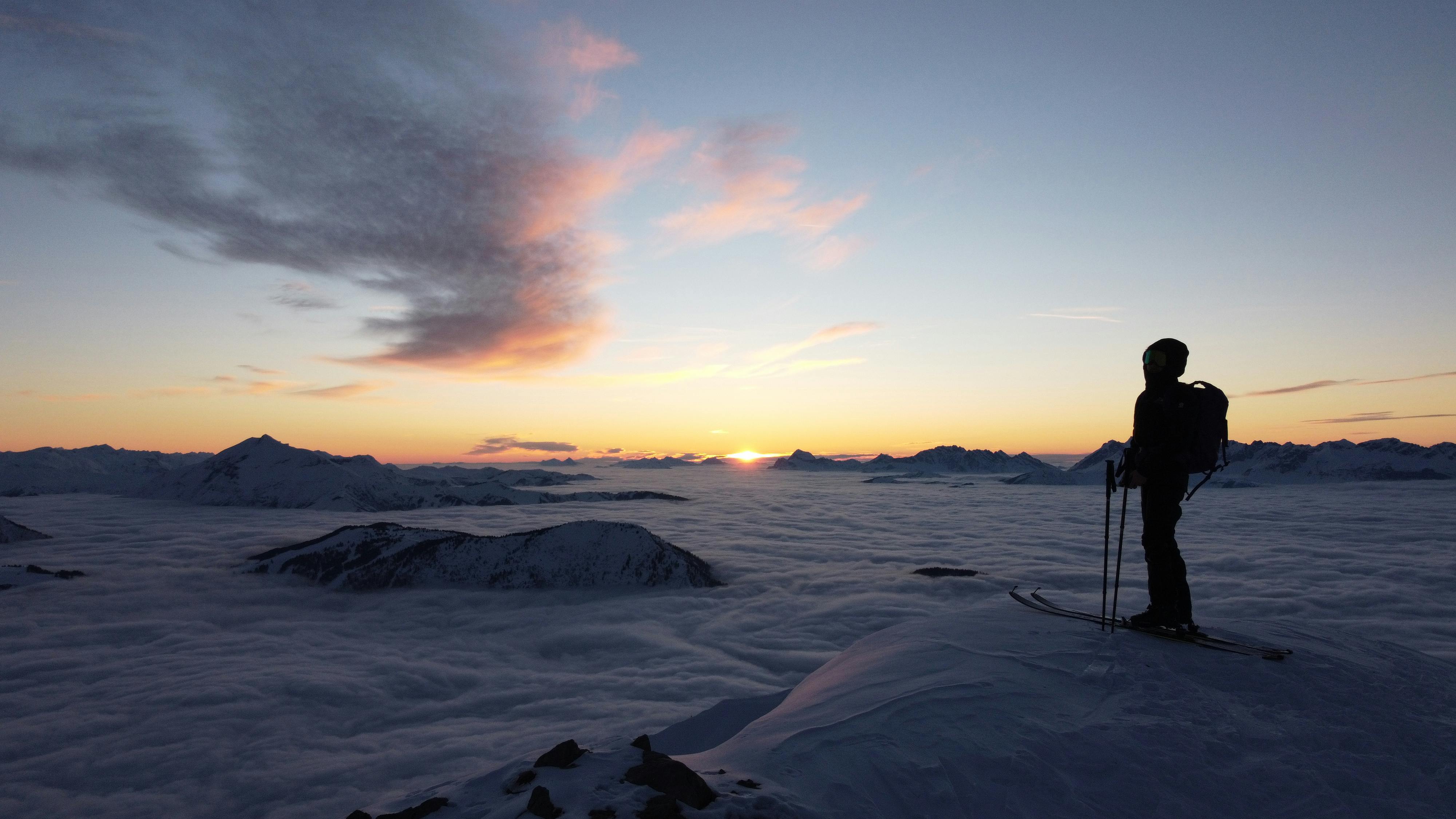 A lone skier stands atop a snowy peak in Les Houches, overlooking a stunning sunset.