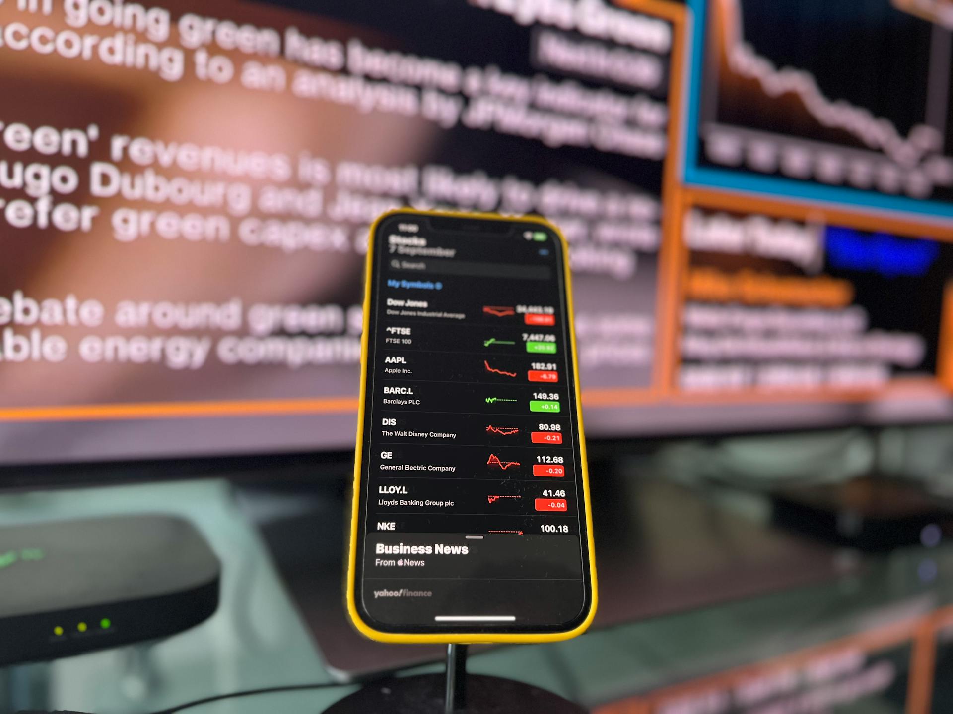 Smartphone showing stock market trends in front of blurred financial news screen indoors.