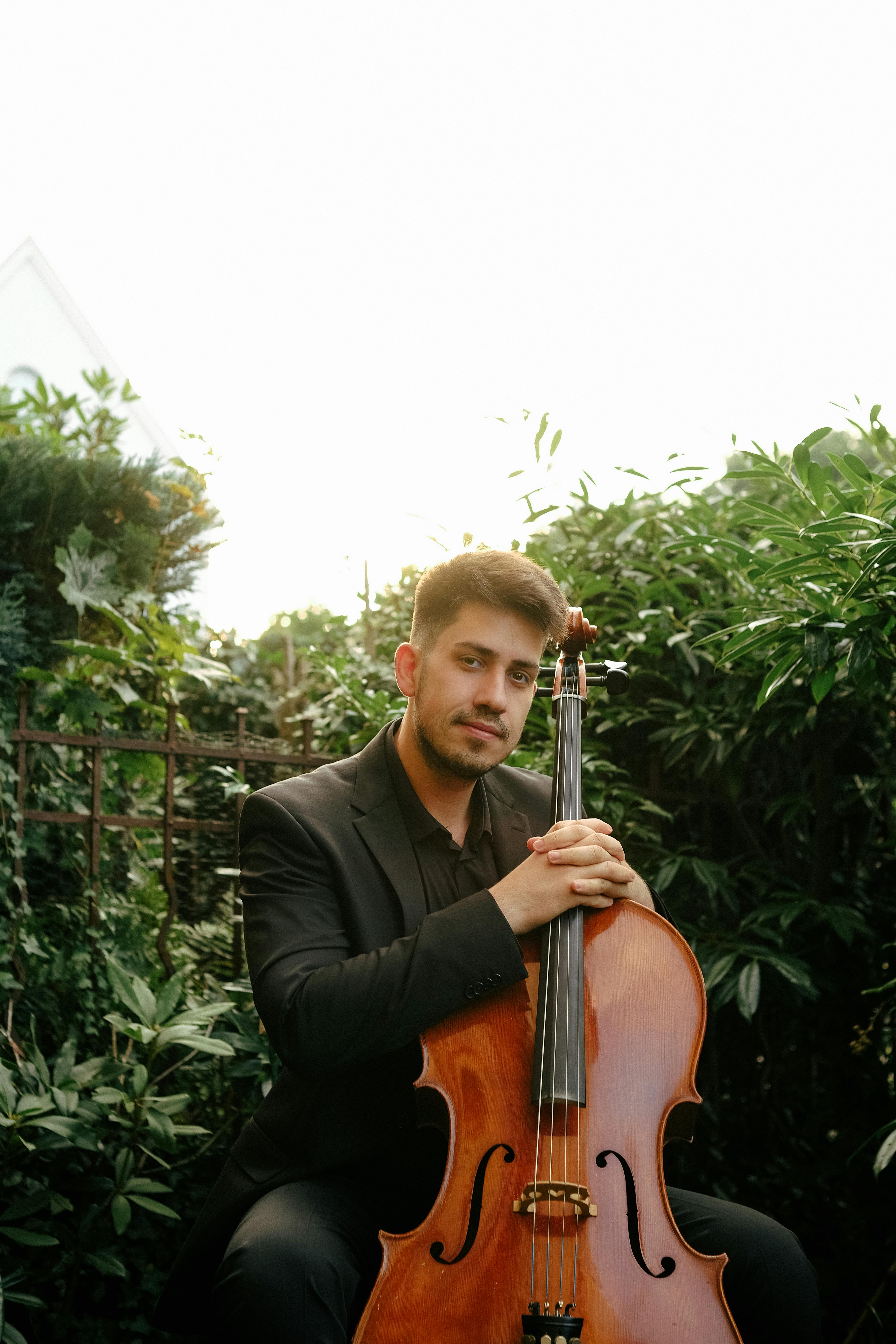 A musician in a black suit poses elegantly with a cello in a garden setting.