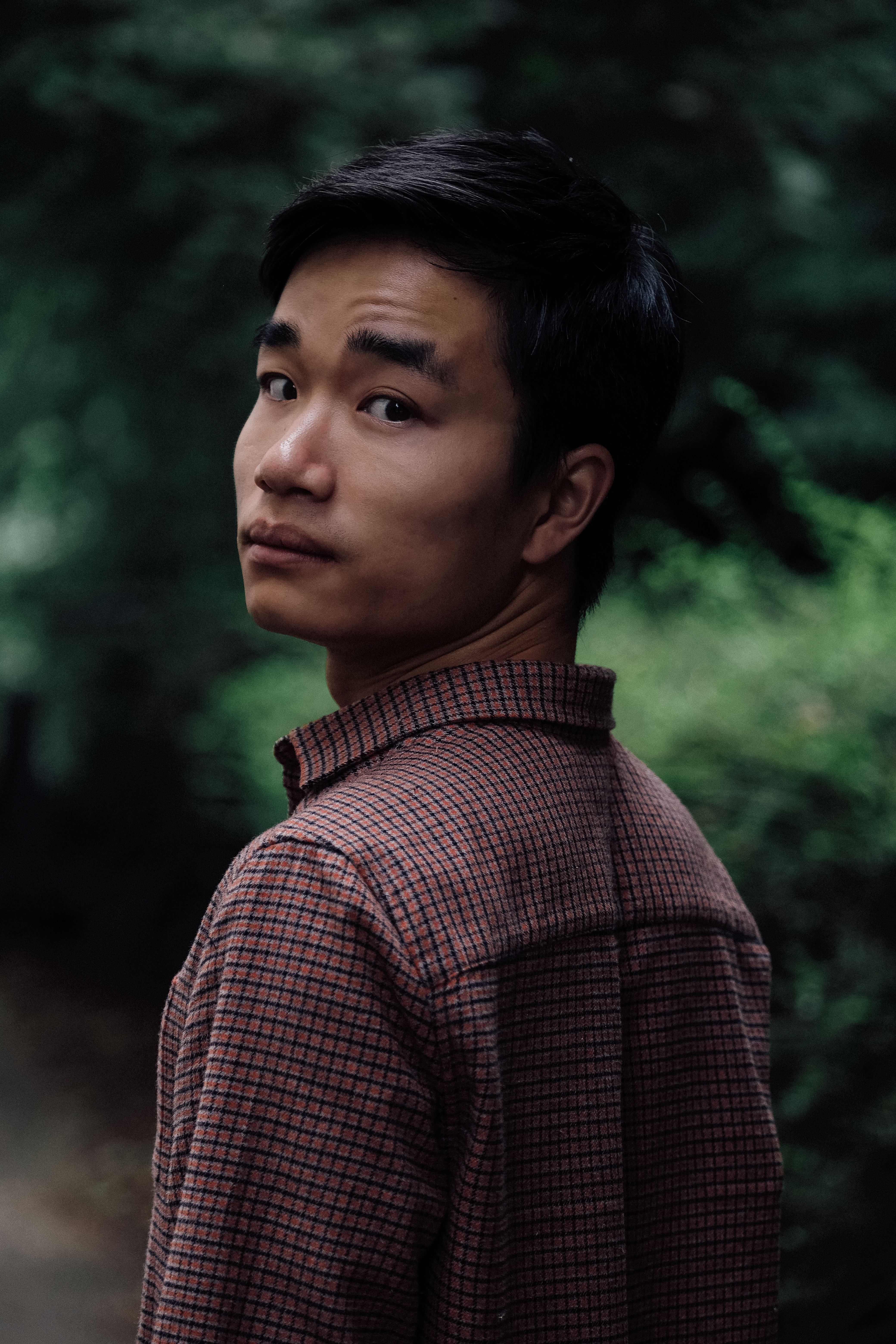 A young Asian man gazes thoughtfully in a forest setting, wearing a checked shirt.