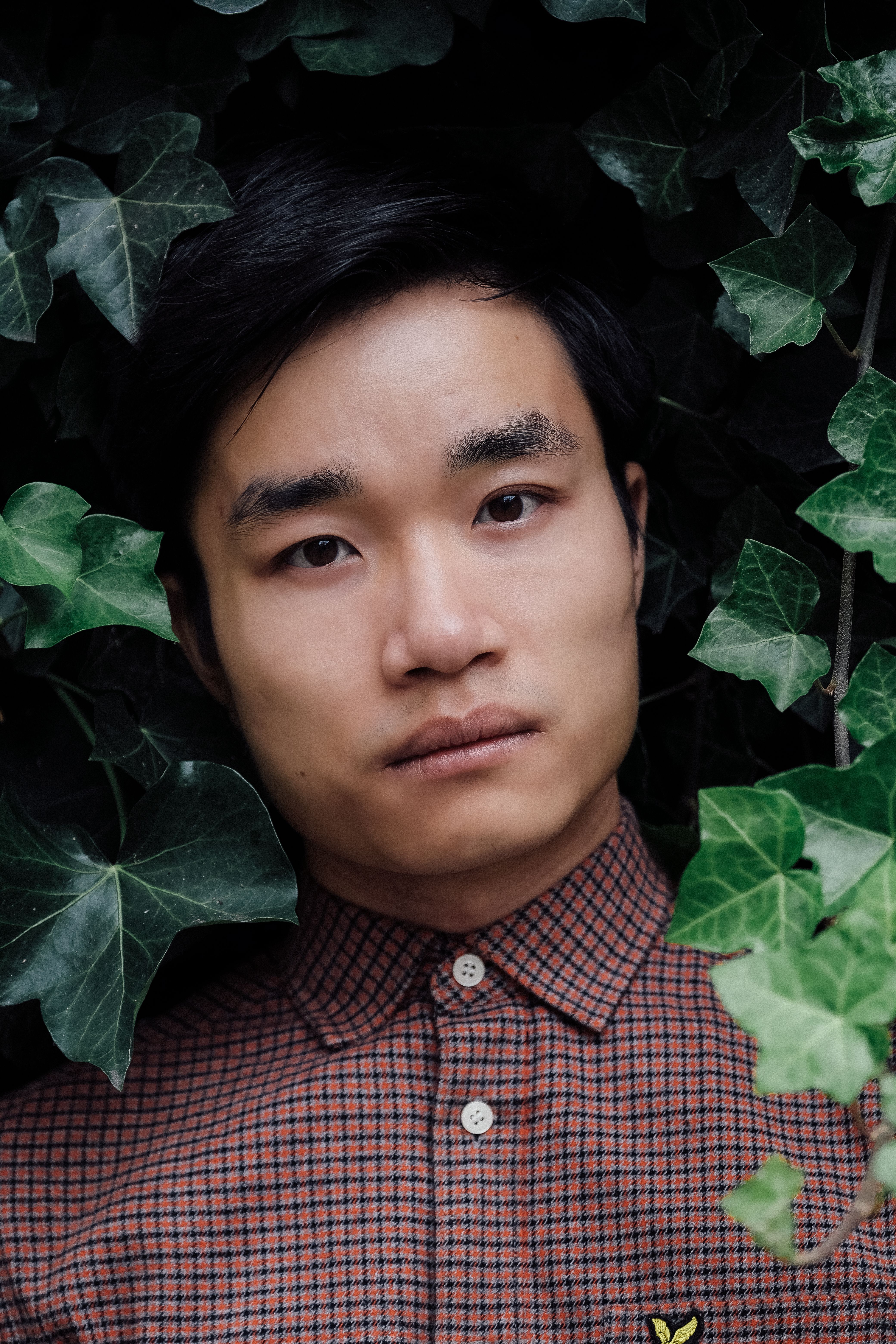 Portrait of a young man surrounded by lush green foliage, wearing a patterned shirt.