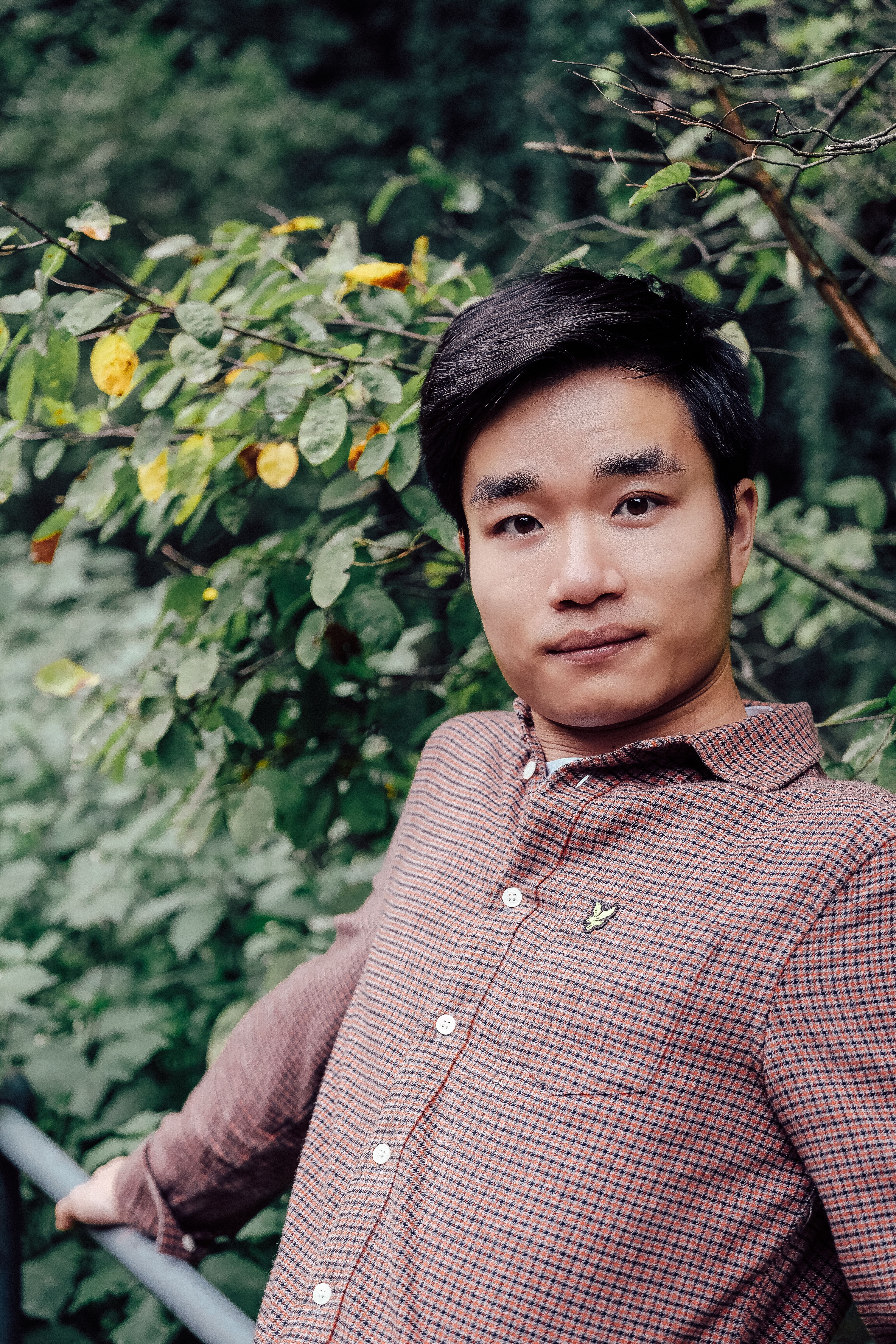 A relaxed portrait of a young man in a forest, surrounded by lush greenery and wearing a checkered shirt.