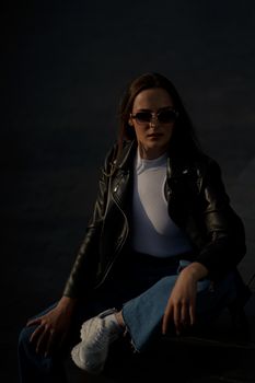 Stylish woman posing in a leather jacket and sunglasses against a dark background.