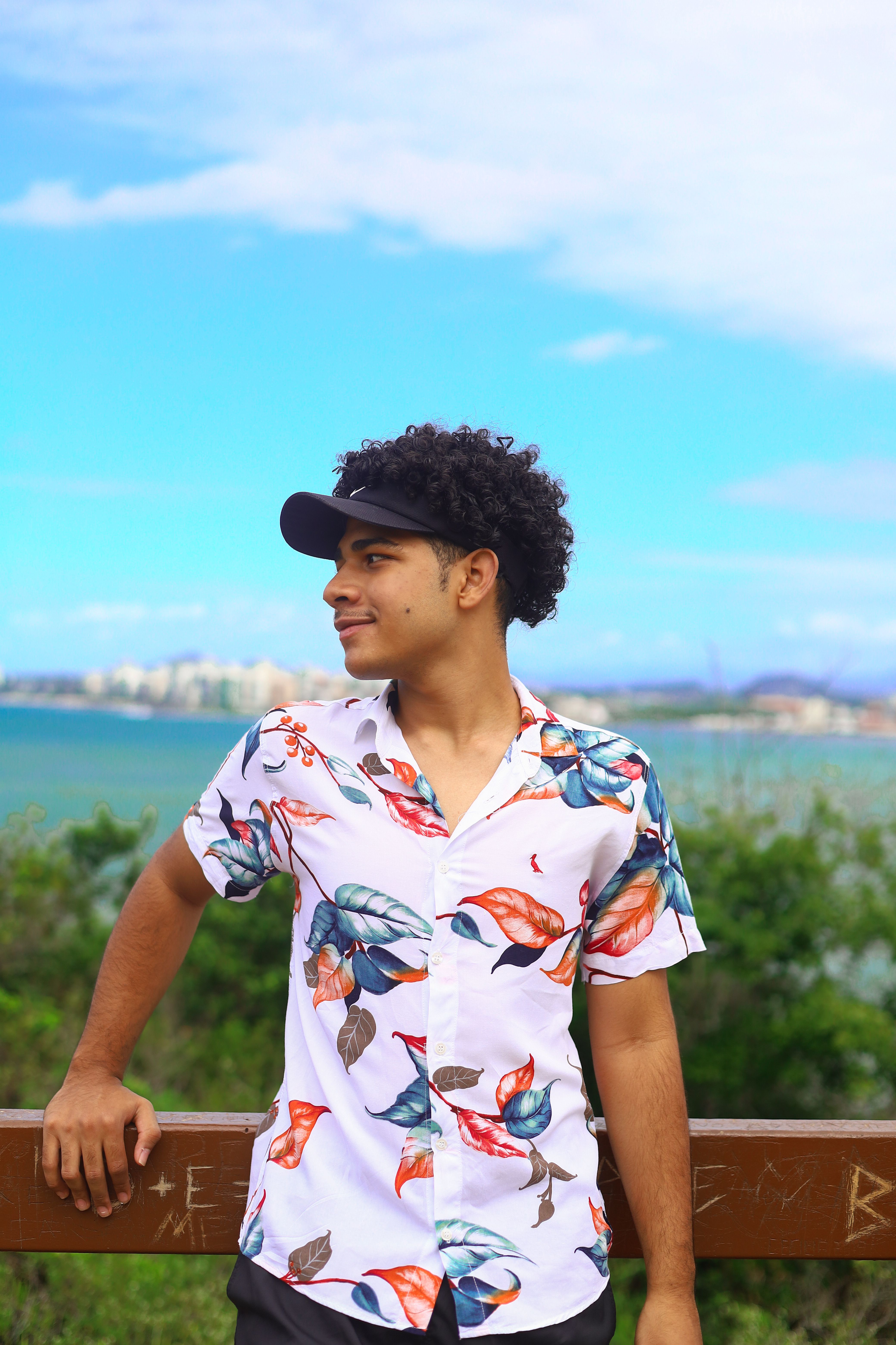 A young man in a floral shirt stands outdoors with a city skyline in the background.