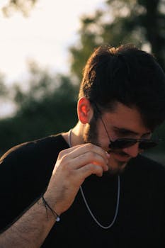 Portrait of a young man adjusting sunglasses outdoors in natural light, with a contemplative demeanor.