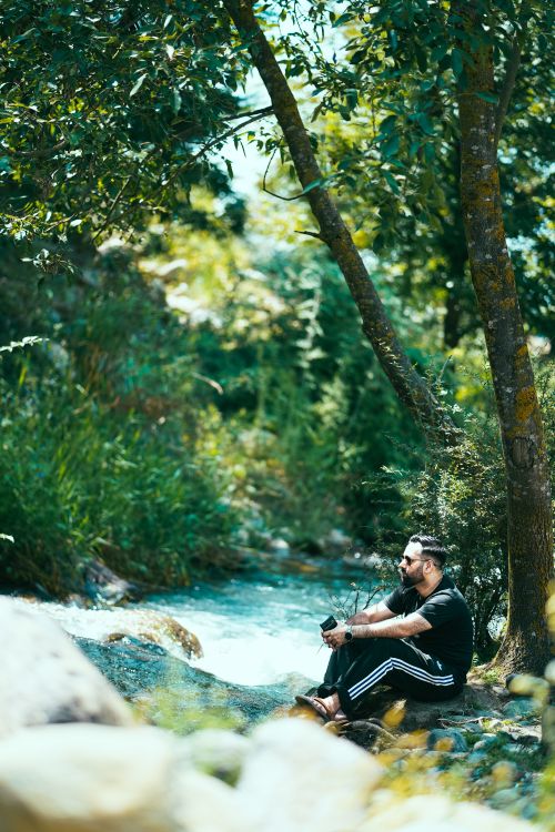 Free A man sits by a tranquil river in a lush forest, enjoying a peaceful summer day. Stock Photo