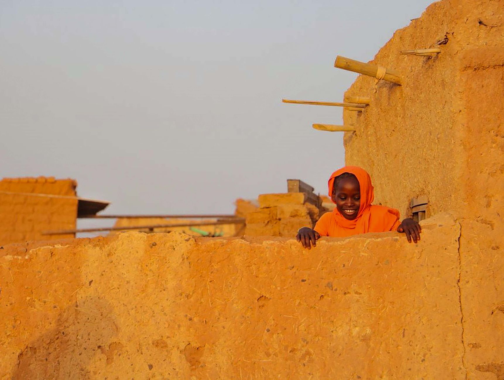A joyful child in bright orange attire smiles over a rustic stone wall in Khartoum, Sudan.