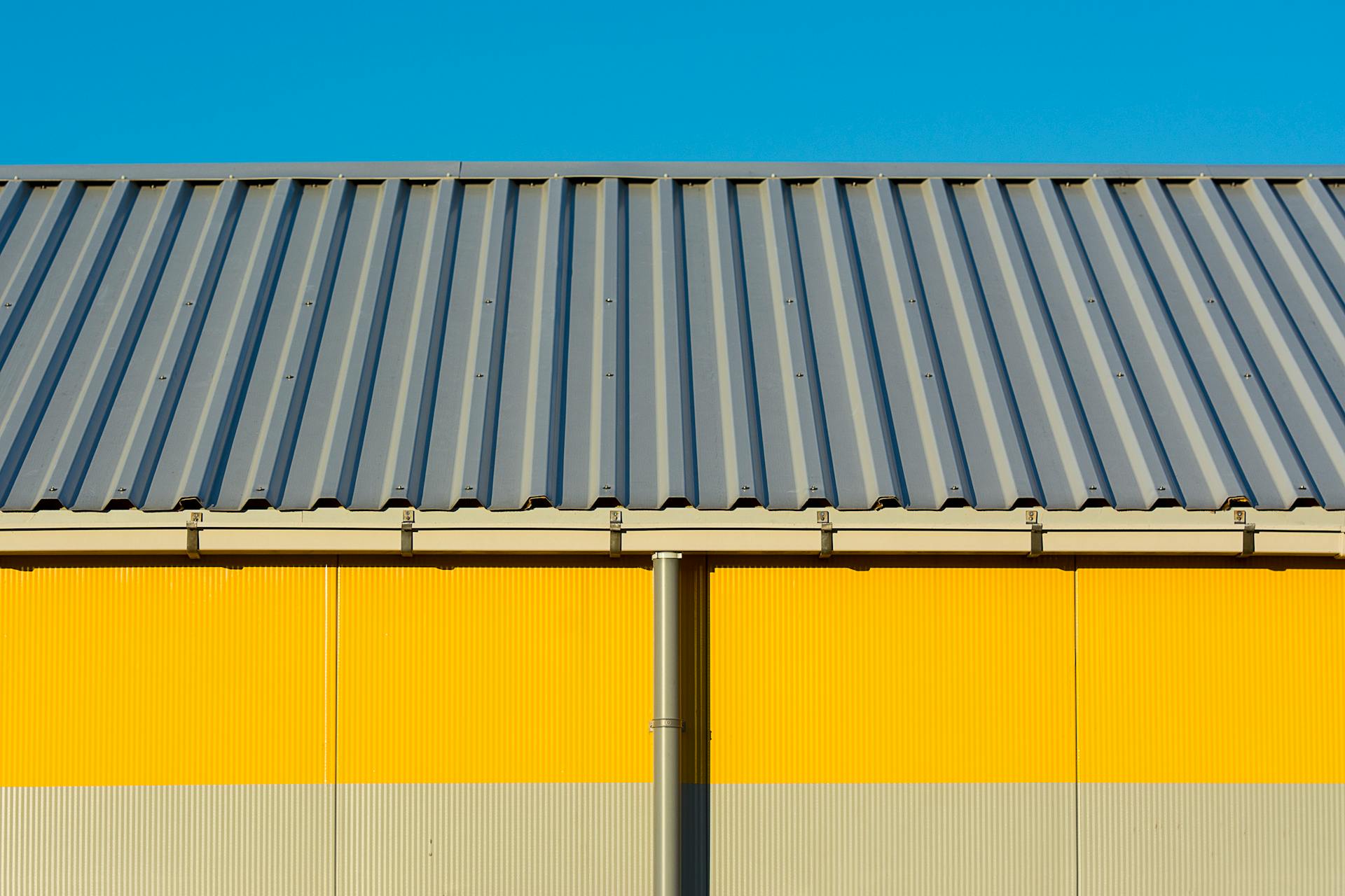 Photo of a yellow warehouse wall with a corrugated metal roof under a clear blue sky.