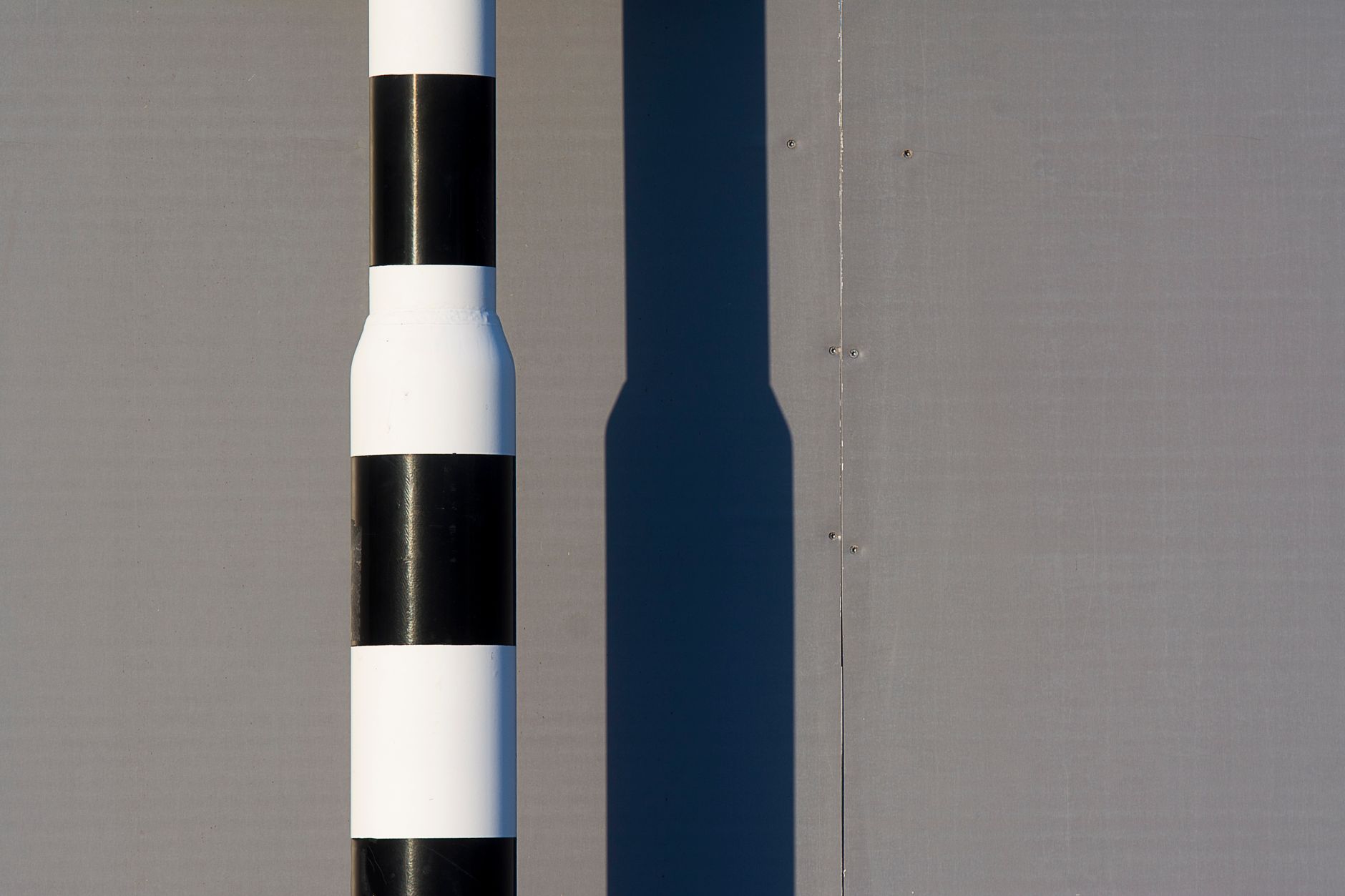 A minimalistic image of a black and white striped post casting a shadow on a gray wall.