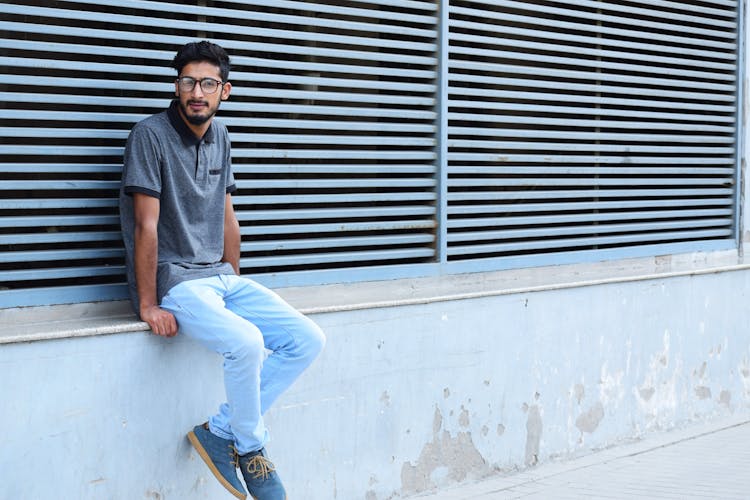 Young Bearded Man In Eyeglasses Sitting On A Wall 